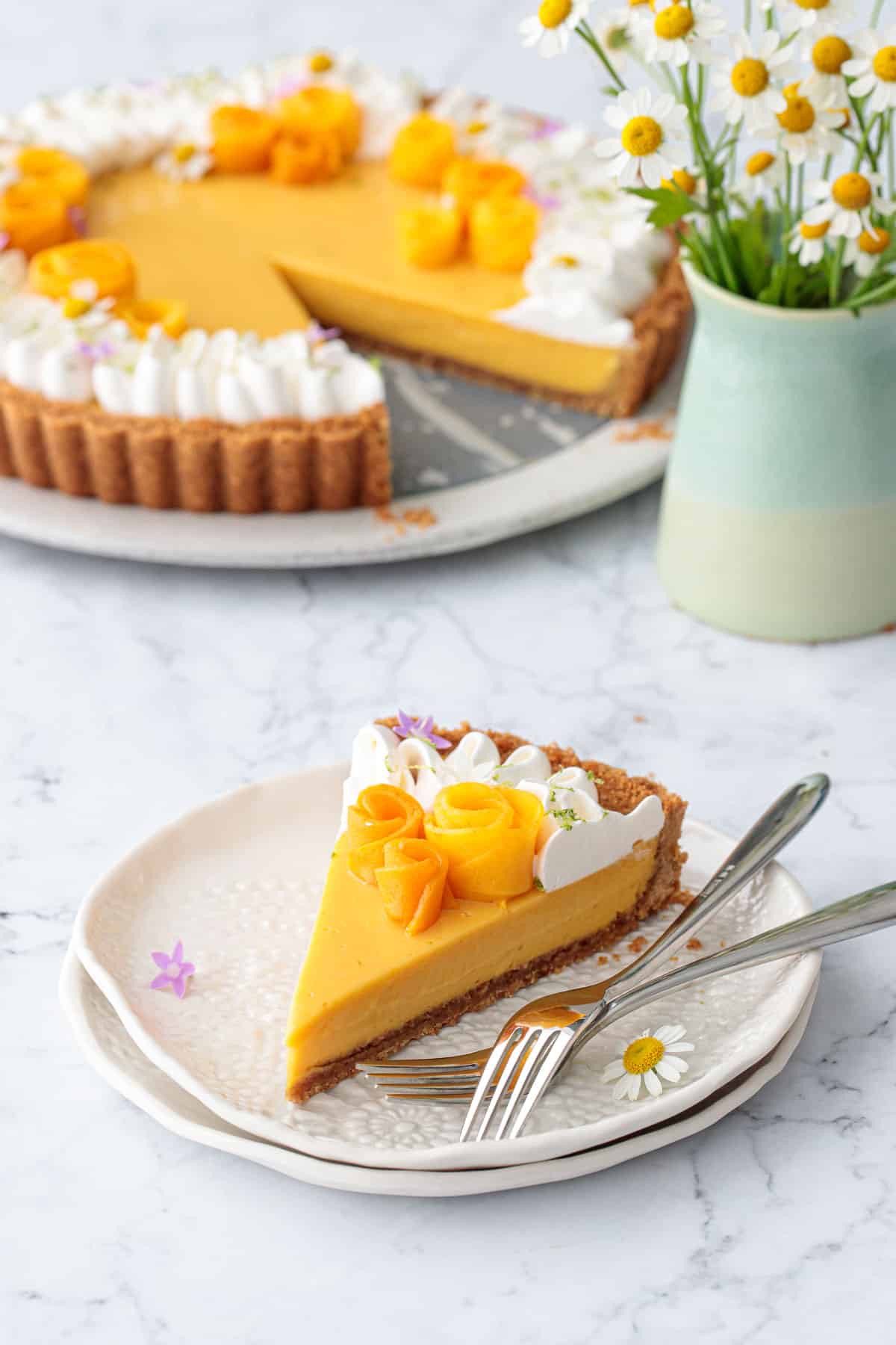Slice of Mango Lime Tart on a white plate with forks, vase of chamomile flowers and the rest of the tart in the background.