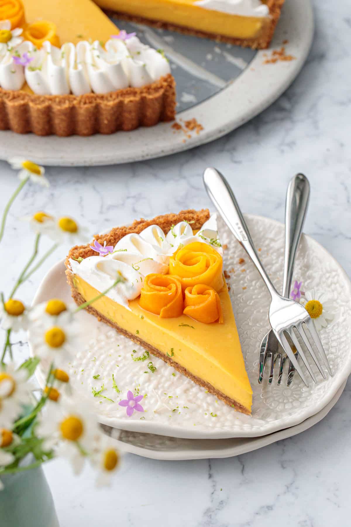Slice of Mango Lime Tart on a plate, chamomile flowers out of focus in the foreground.