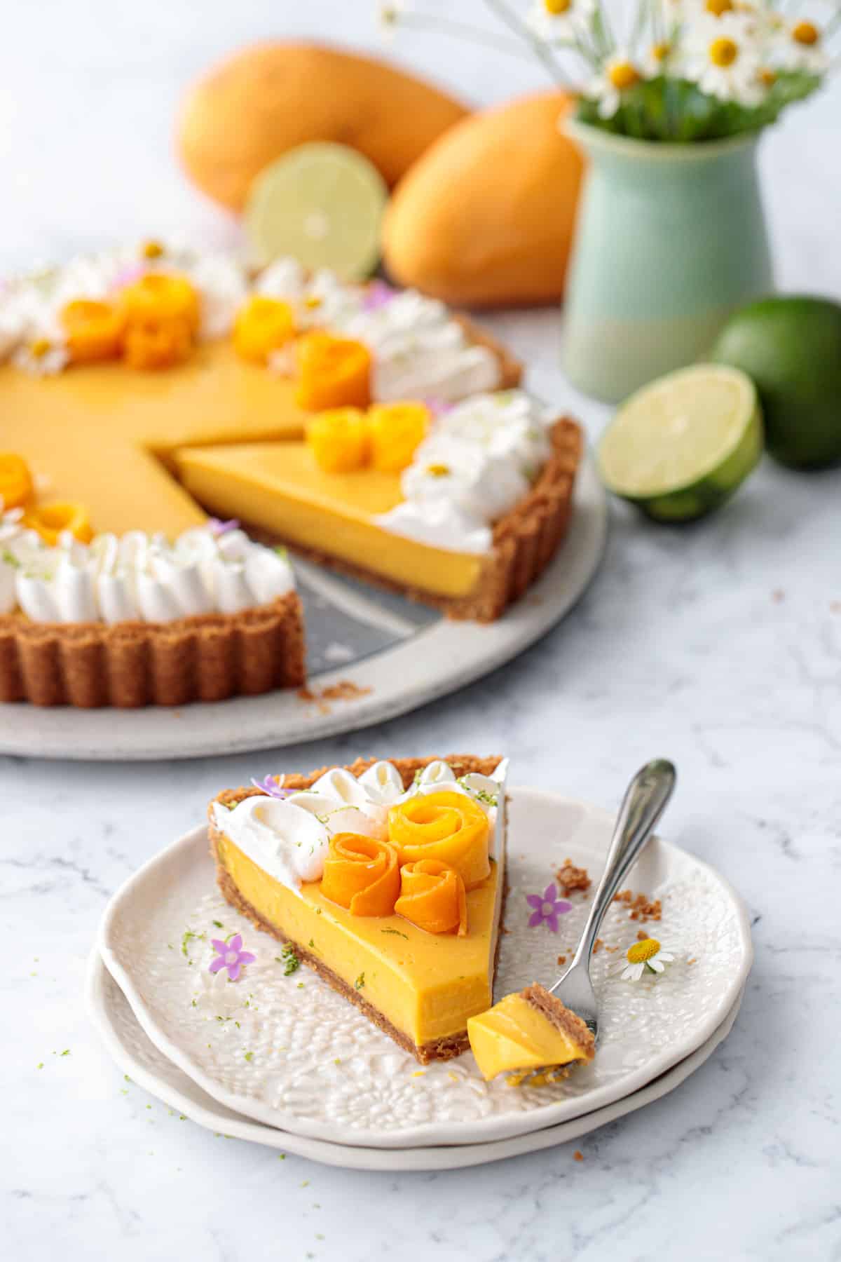 Slice of Mango Lime Tart on a white plate, with the full tart, vase of flowers, plus whole mangos and limes in the background.