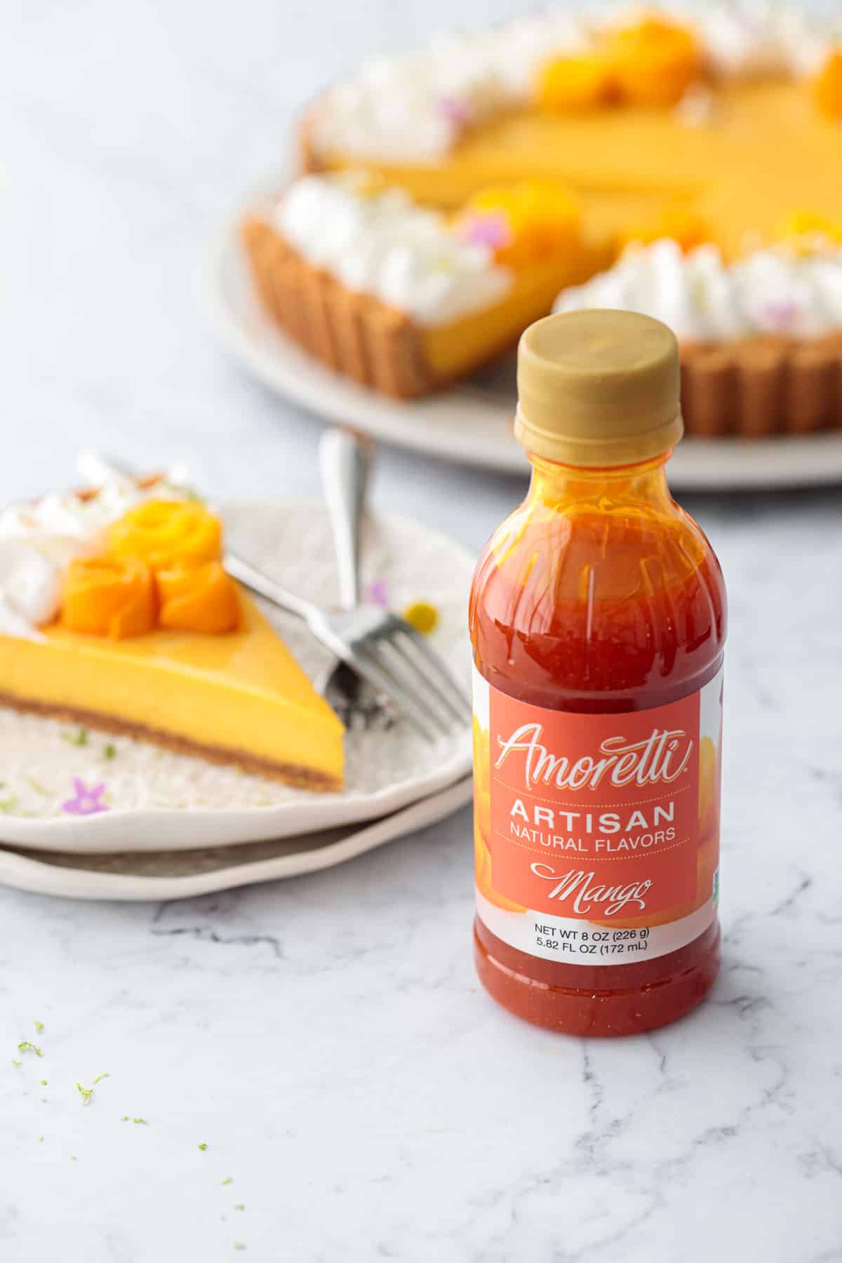 Bottle of Amoretti Natural Artisan Mango Flavor with a slice of Mango Lime Tart on a plate and the full tart out of focus in the background.