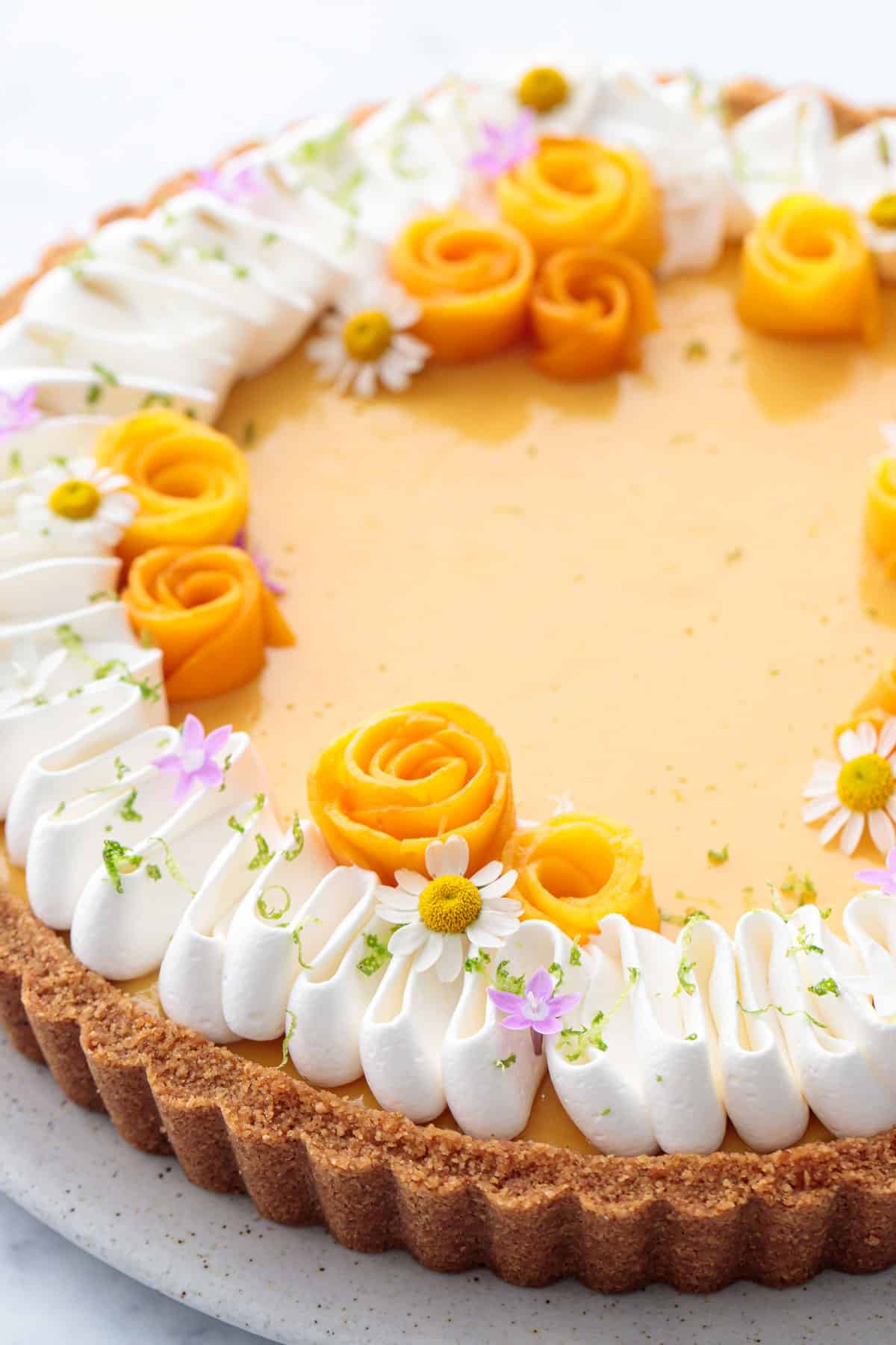 Closeup of a bright yellow Mango Lime Tart topped with mango flowers, purple and white edible flowers, and a squiggle of qhipped cream with lime zest.