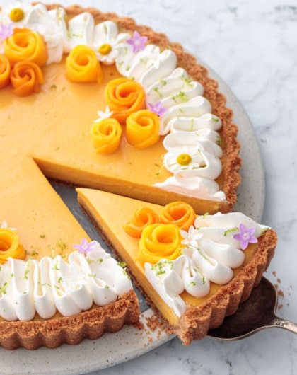 Mango Lime Tart with a slice cut out of it to show the creamy texture inside, decorated with whipped cream and edible flowers.