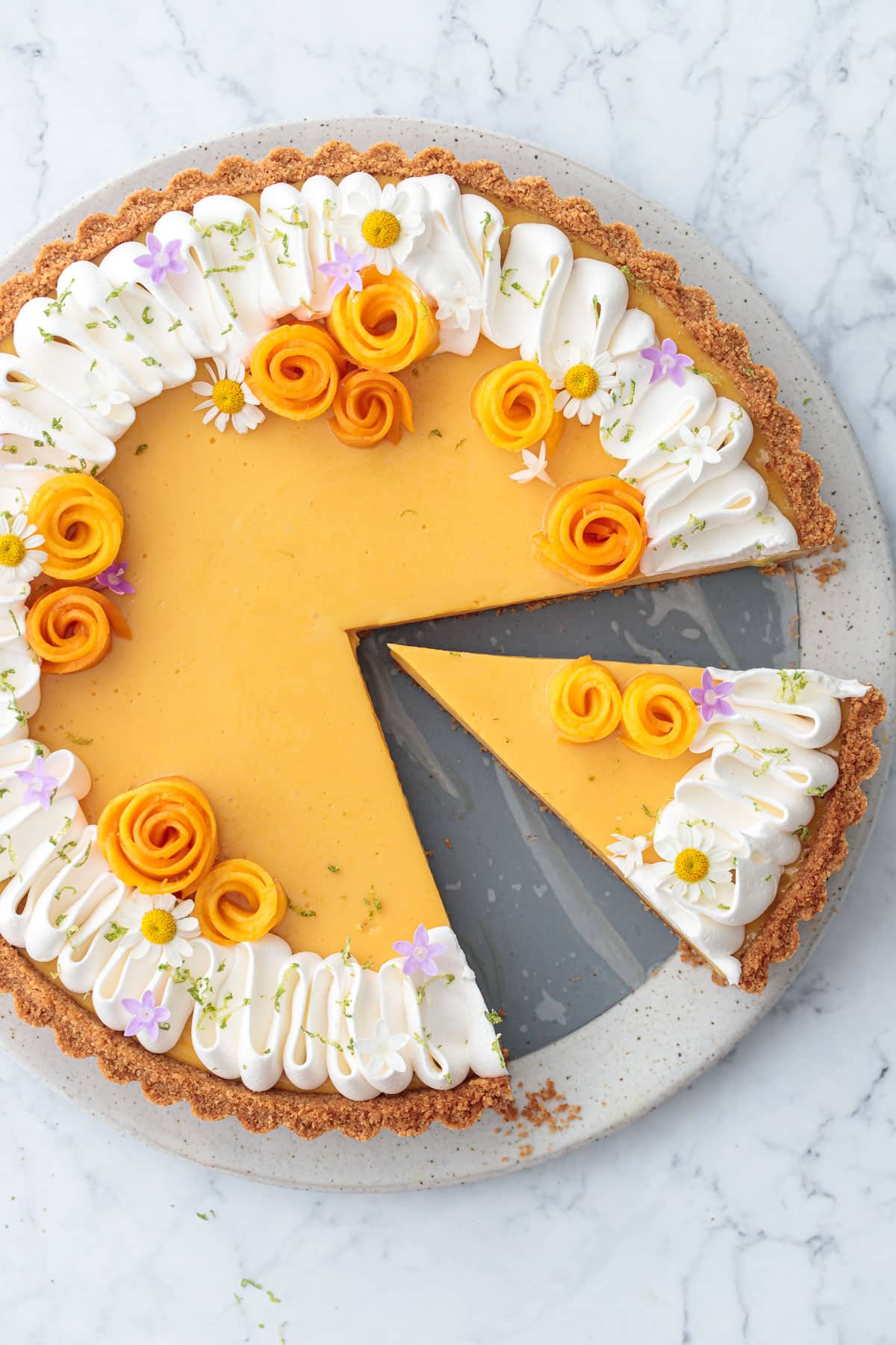 Overhead, Mango Lime Tart with a slice taken out of it, topped with whipped cream, lime zest, and edible flowers.