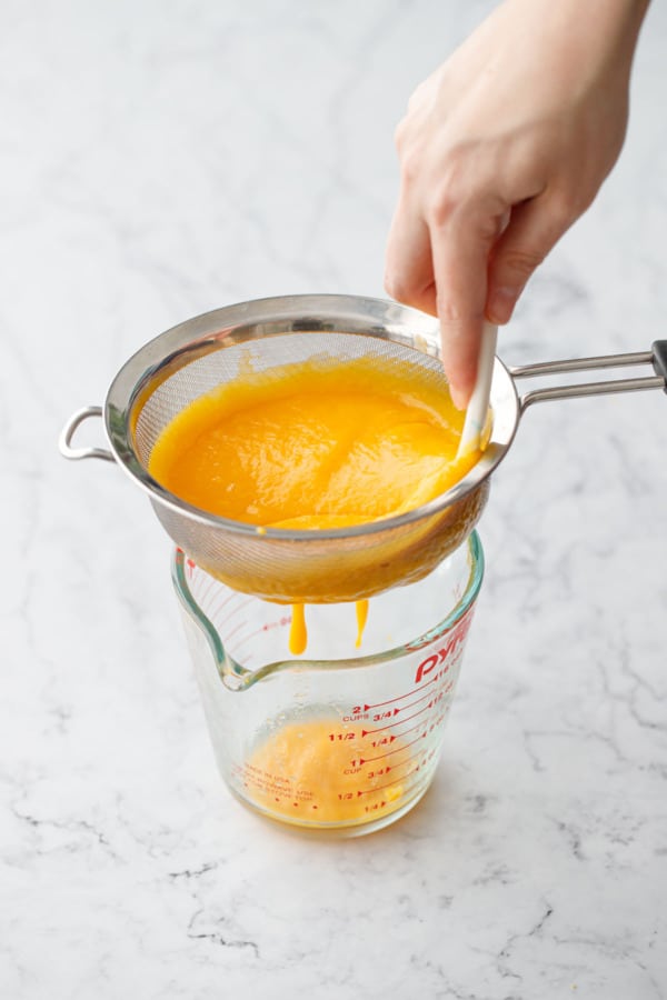 Pressing mango puree through a fine mesh sieve set over a glass measuring cup.