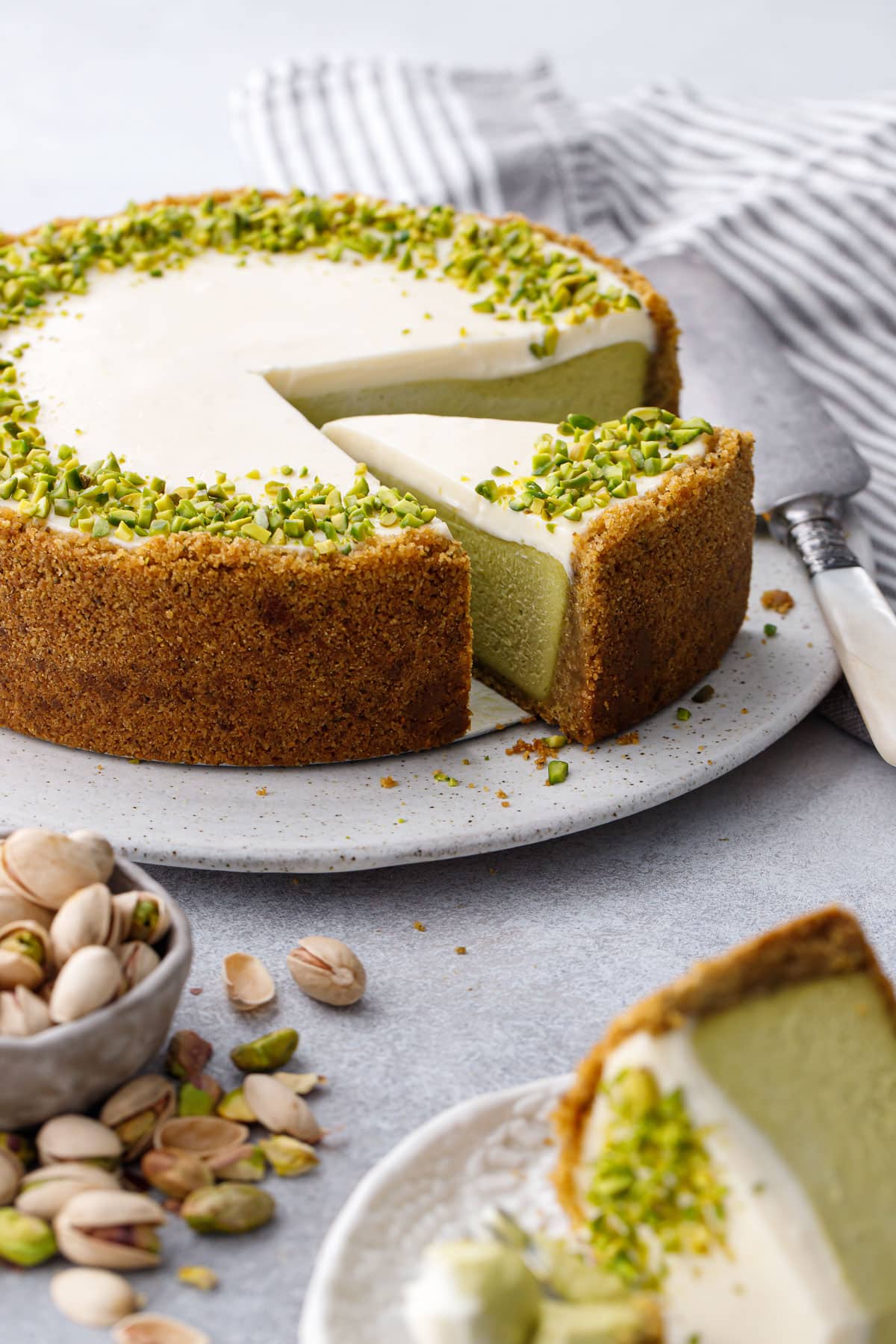 Slice out of a full Pistachio Sour Cream Cheesecake, with out of focus bowl of pistachios and cheesecake slice in the foreground.