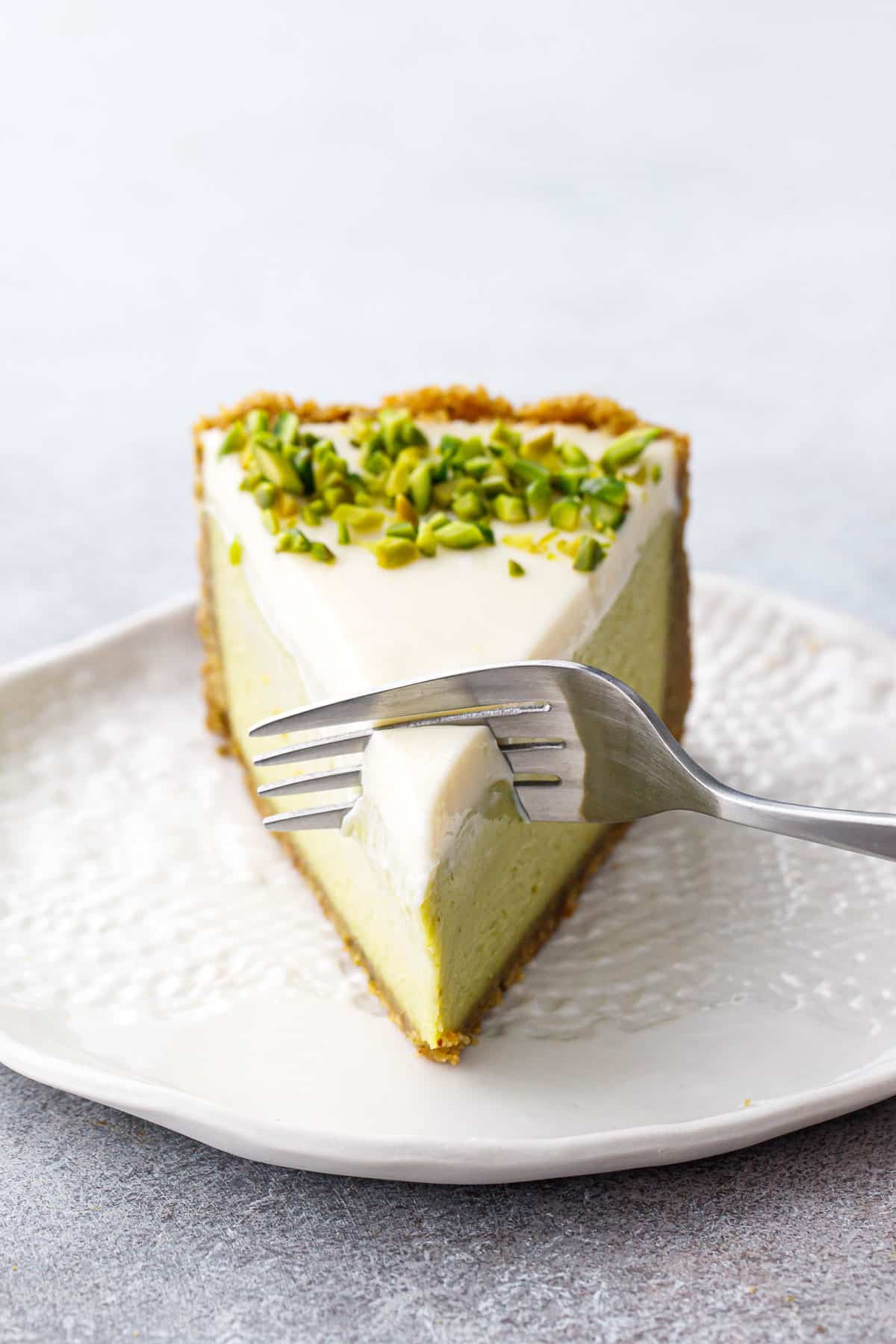 Triangular slice of Pistachio Sour Cream Cheesecake on a white plate, with a fork cutting through the tip of the slice to show the creamy texture.