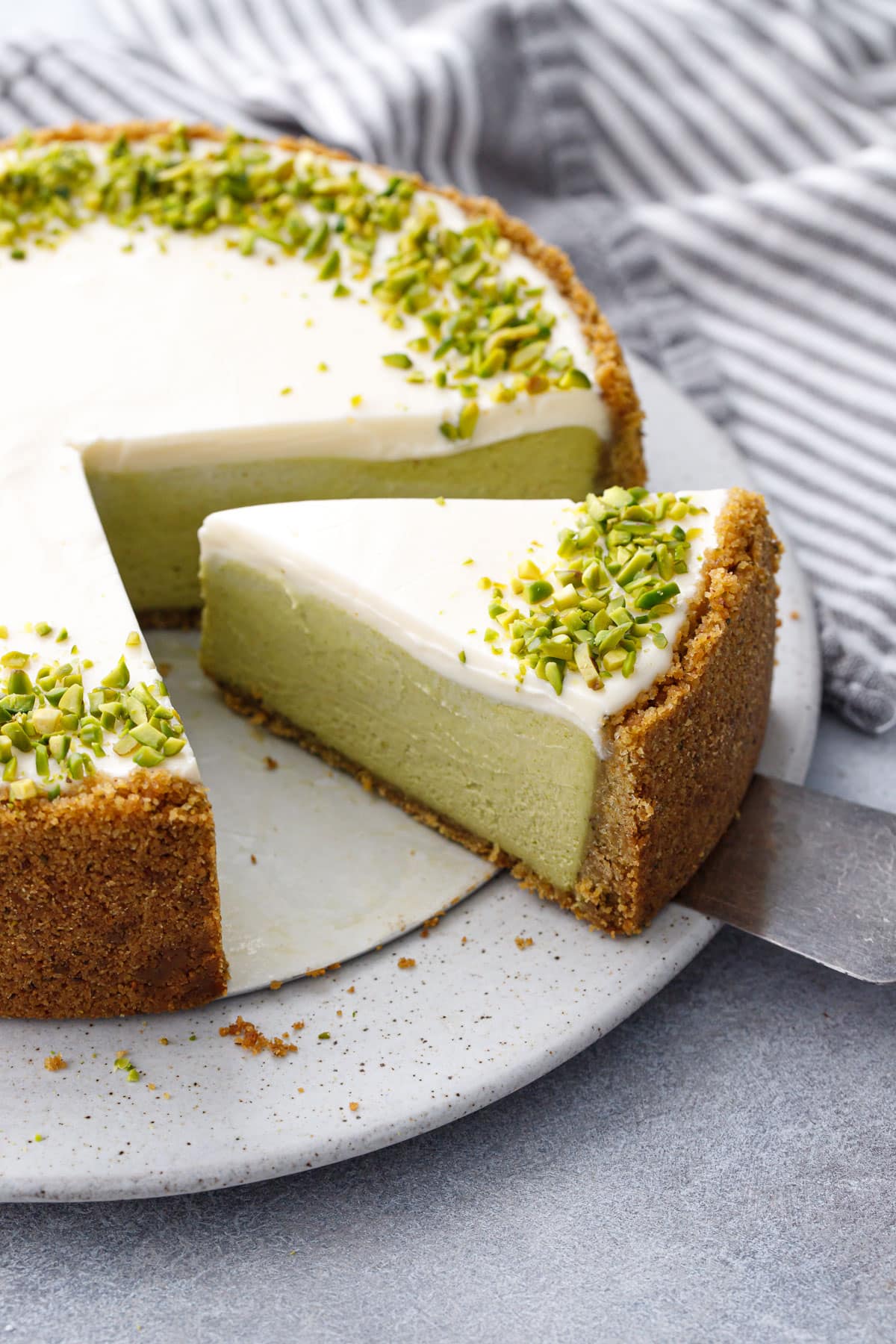 Slice of Pistachio Sour Cream Cheesecake on a cake plate with cake server, topped with a layer of white sour cream and a border of green chopped pistachios.