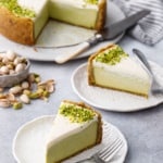 Slices of Pistachio Sour Cream Cheesecake on white porcelain plates and silver forks, full cheesecake with slices cut out on the background with a bowl of scattered pistachios.