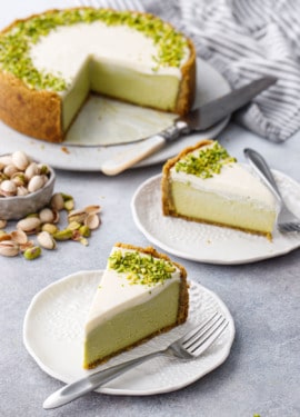 Slices of Pistachio Sour Cream Cheesecake on white porcelain plates and silver forks, full cheesecake with slices cut out on the background with a bowl of scattered pistachios.