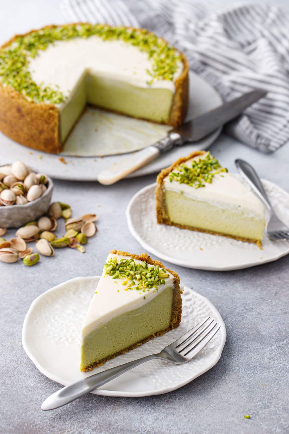 Slices of Pistachio Sour Cream Cheesecake on white porcelain plates and silver forks, full cheesecake with slices cut out on the background with a bowl of scattered pistachios.