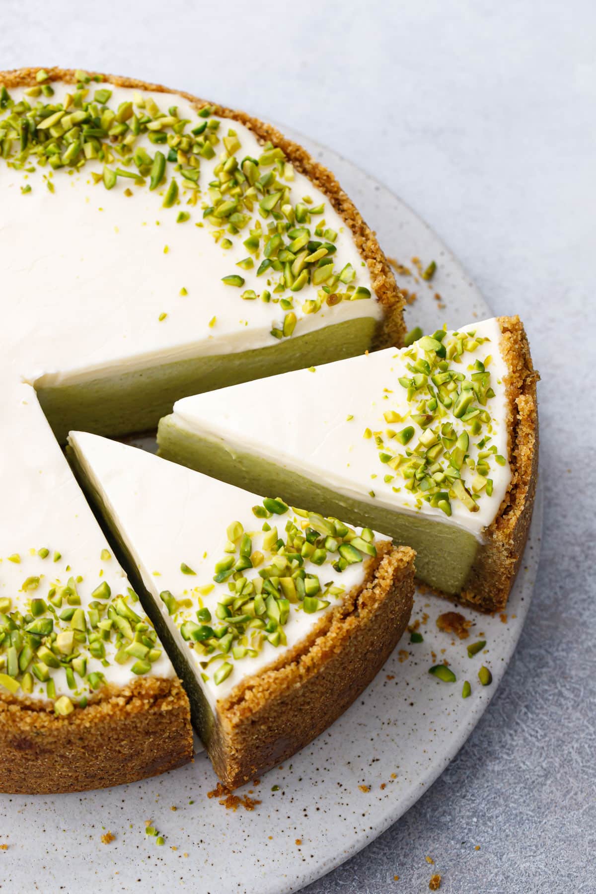 Two slices out of a full round Pistachio Sour Cream Cheesecake, with a white sour cream topping and a border of chopped green pistachios.