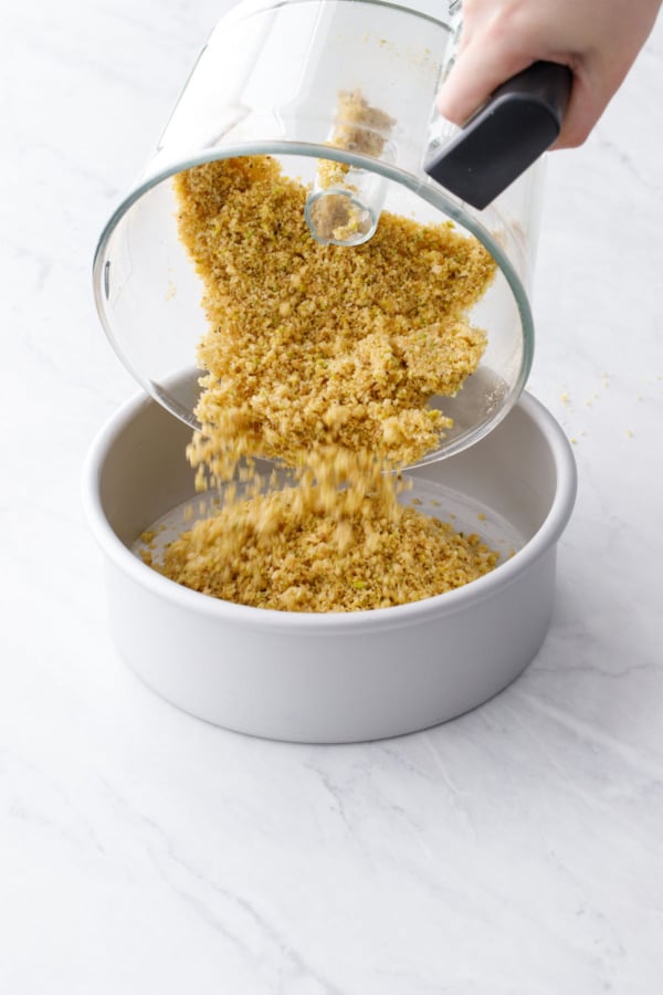 Pouring crushed vanilla wafer cookies and pistachios into a cheesecake pan to make the crust.