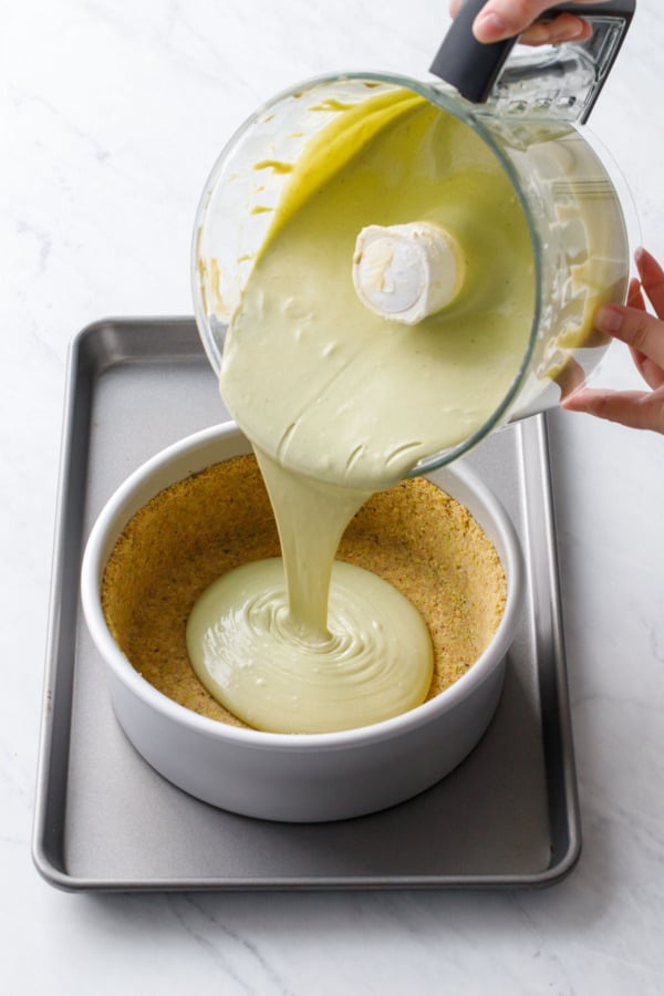 Pouring the green pistachio cheesecake filling from a food processor into the pre-baked crust.