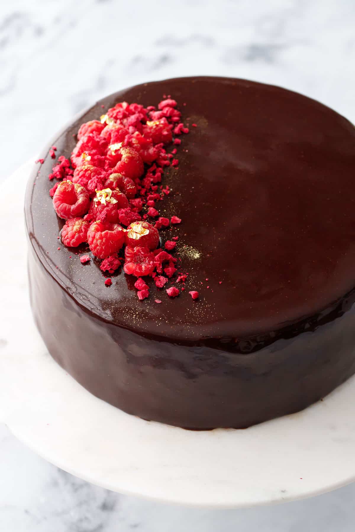 Chocolate Raspberry Mousse Cake with a super shiny chocolate mirror glaze and decorated with fresh and freeze dried raspberries and gold leaf accents.