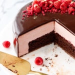 Raspberry and chocolate entremet-style cake with a dark chocolate mirror glaze with a few slices cut out to show the interior layers, with two raspberries and a gold cake server on the side.