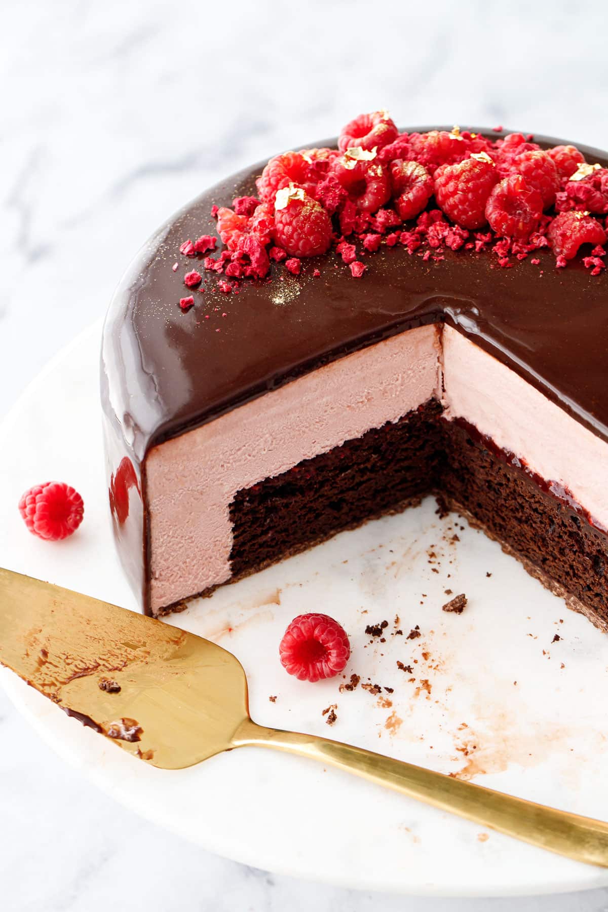 Raspberry and chocolate entremet-style cake with a dark chocolate mirror glaze with a few slices cut out to show the interior layers, with two raspberries and a gold cake server on the side.