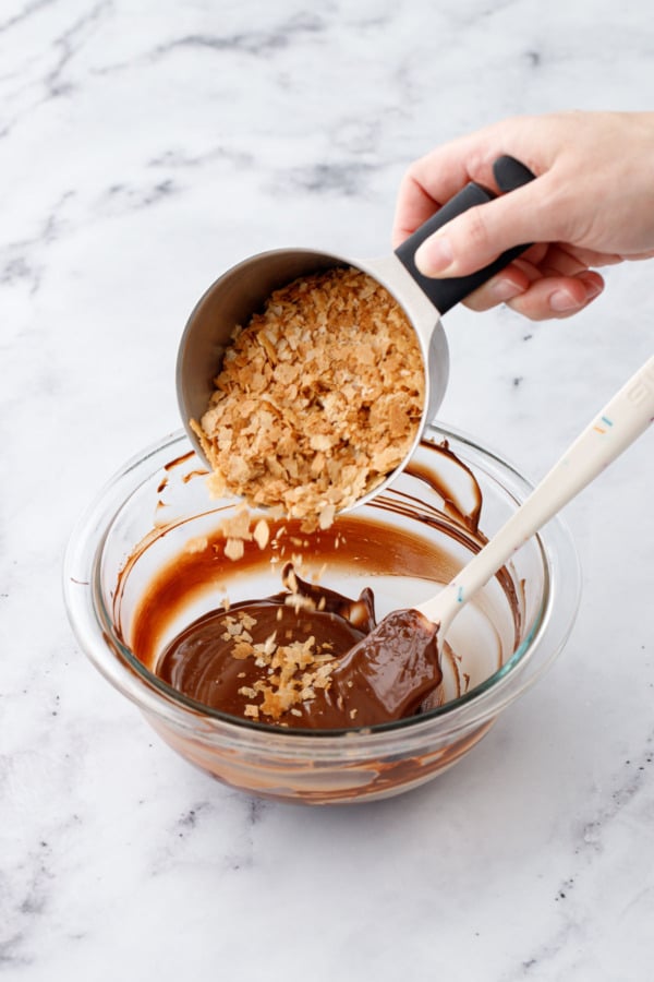 Cup of feuilletine pouring into a bowl of melted chocolate mixed with nutella.