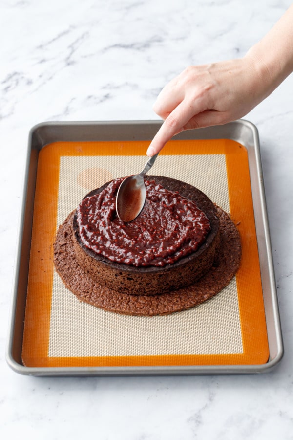 Spoon spreading a layer of chocolate raspberry jam on top of the chocolate cake.