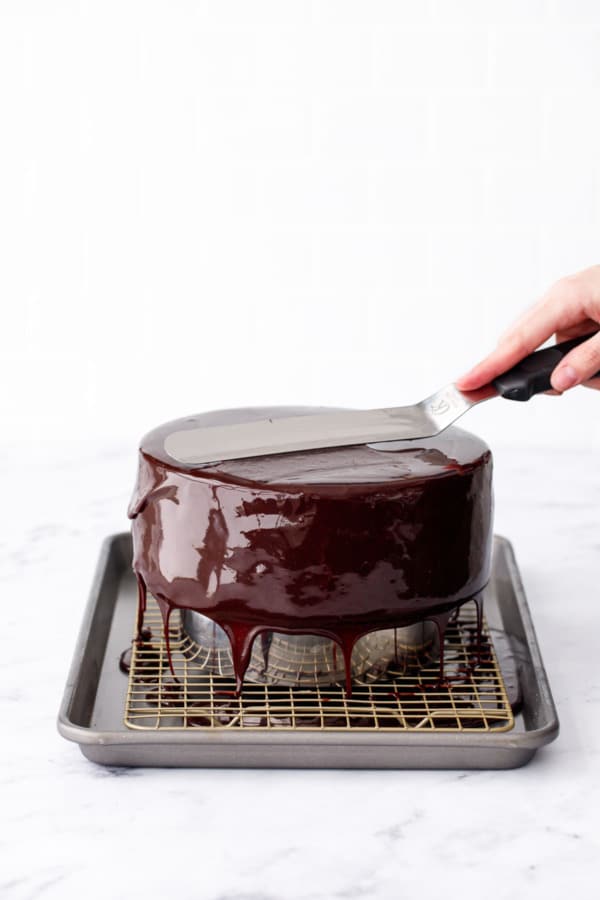 Offset spatula leveling off the top of the dark chocolate mirror-glazed cake, excess dripping down the sides into the baking sheet below.