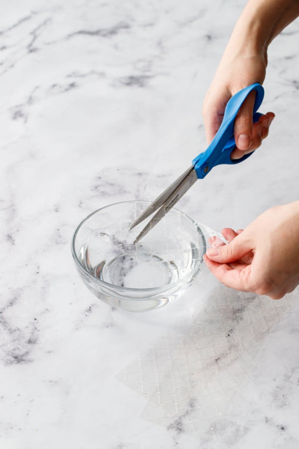 Scissors cutting sheets of leaf gelatin into a bowl of cold water to soften.