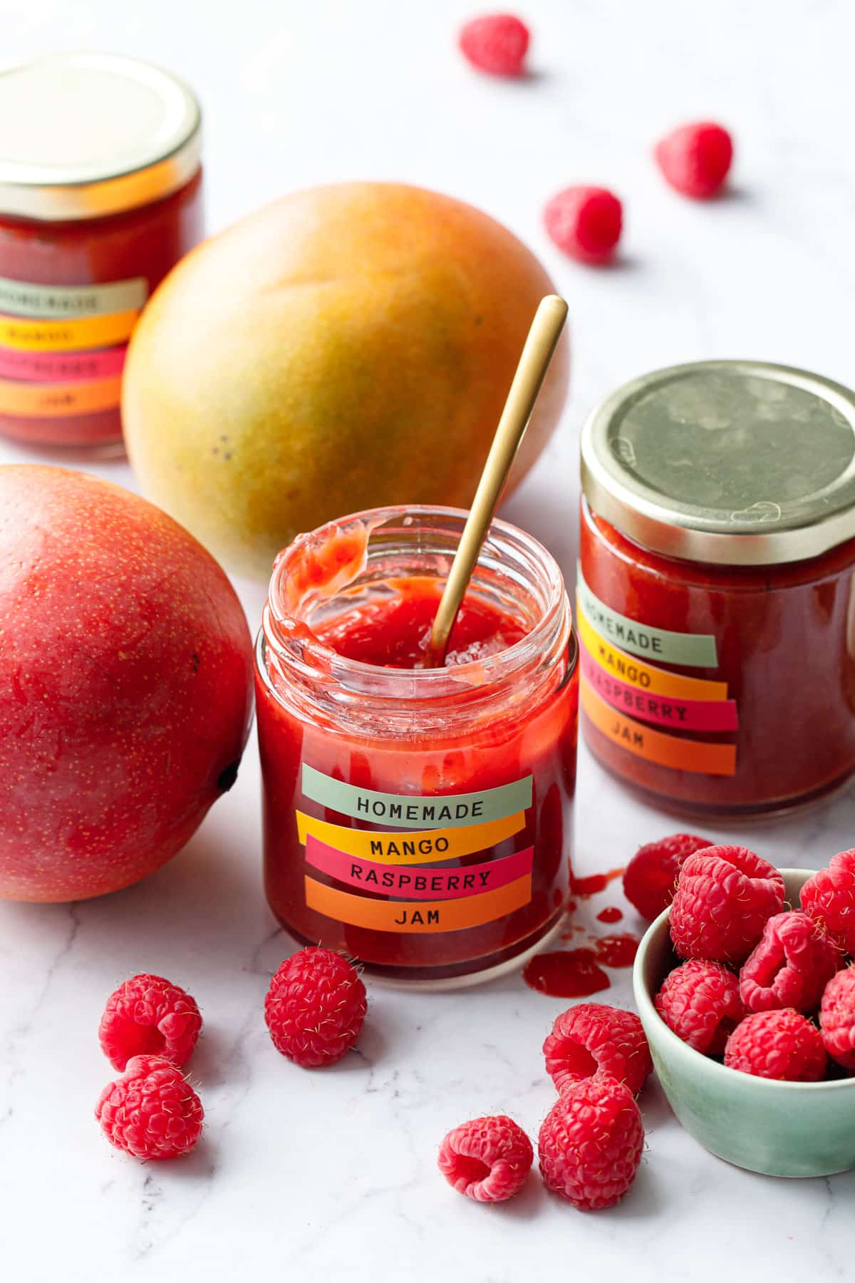 Jars of Mango Raspberry Jam, one open to show the texture of the jam, with scattered mango fruits and raspberries around.