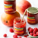 Jars of Mango Raspberry Jam with colorful label design, with bowl of raspberries and fresh mangoes scattered around.