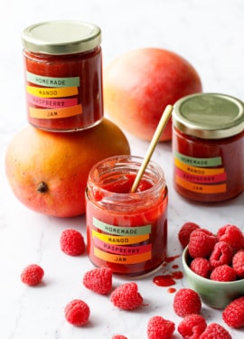 Jars of Mango Raspberry Jam with colorful label design, with bowl of raspberries and fresh mangoes scattered around.
