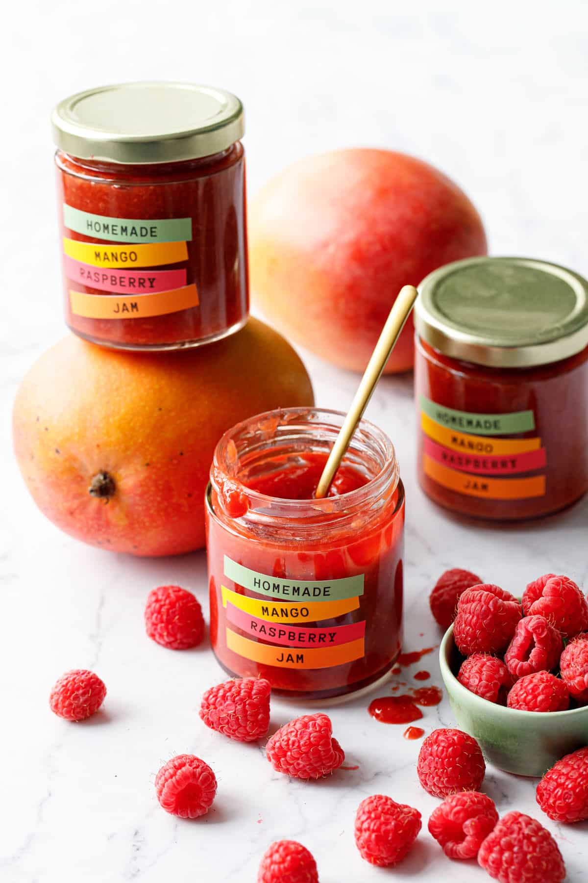 Jars of Mango Raspberry Jam with colorful label design, with bowl of raspberries and fresh mangoes scattered around.