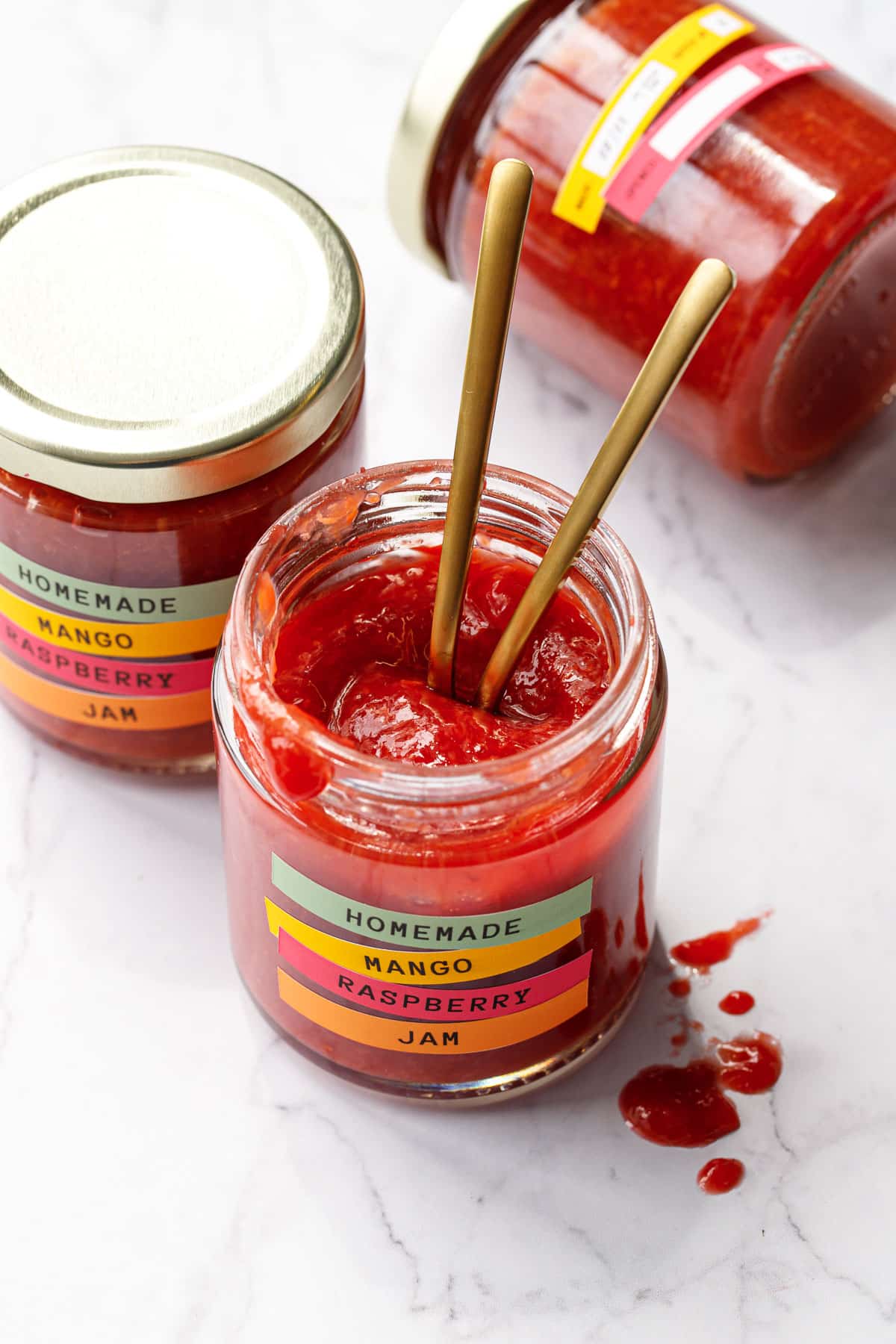 Open jar of Mango Raspberry Jam with two gold spoons down inside, two more jars behind and on the side.