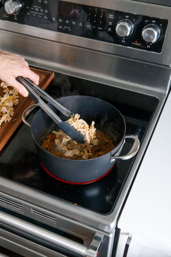 Transferring shredded chicken thighs with tongs back to saucepan with reduced sauce mixture.