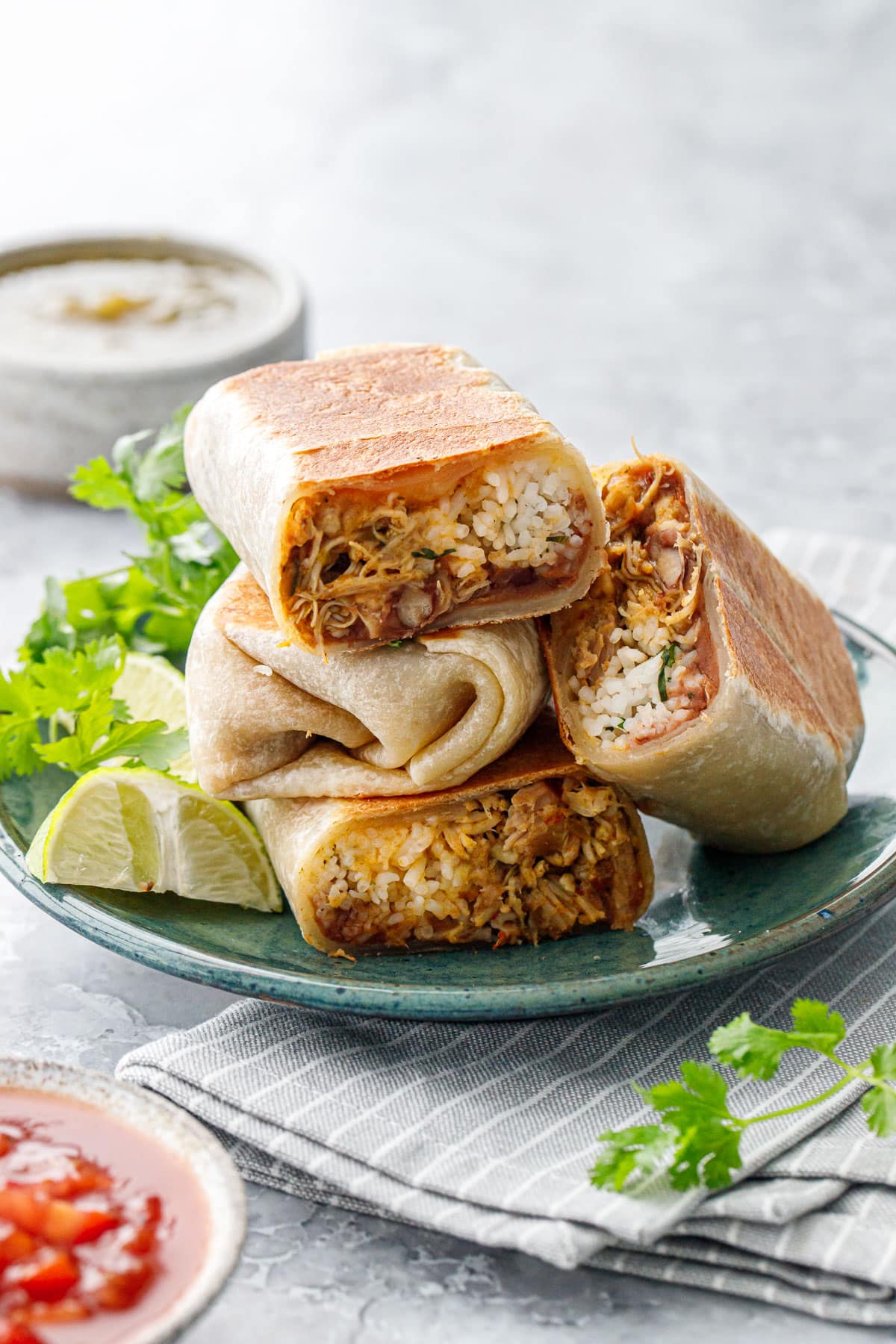 Crispy Chicken, Rice, & Bean Burritos stacked on a plate with cilantro and limes, with gray stripe napkin, and bowls of red and green salsa in the foreground and background.