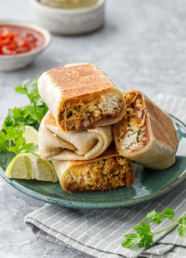 Crispy Chicken, Rice, & Bean Burritos stacked on a green ceramic plate, some burritos cut in half to show the filling side with cilantro, limes, and bowl of salsa on the side.