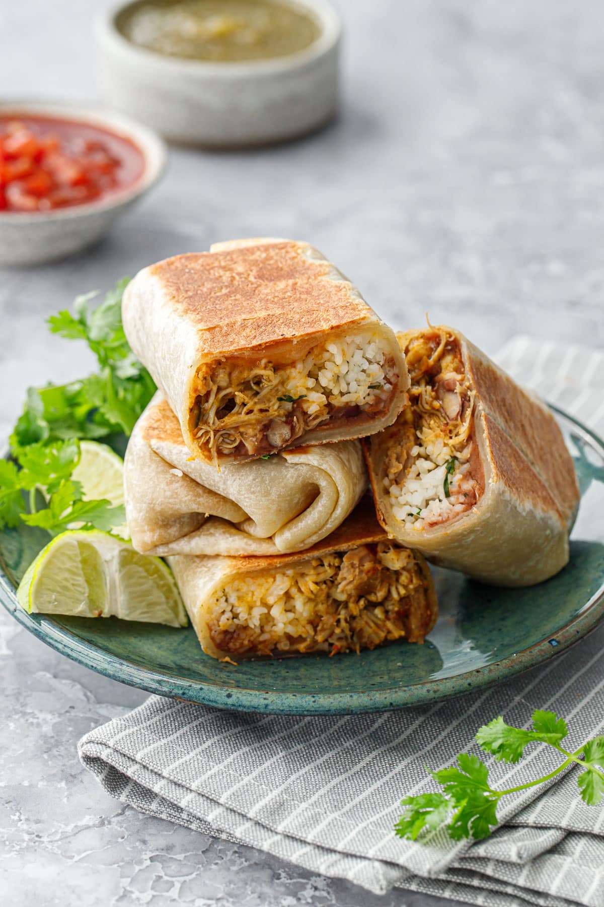 Crispy Chicken, Rice, & Bean Burritos stacked on a green ceramic plate, some burritos cut in half to show the filling side with cilantro, limes, and bowl of salsa on the side.