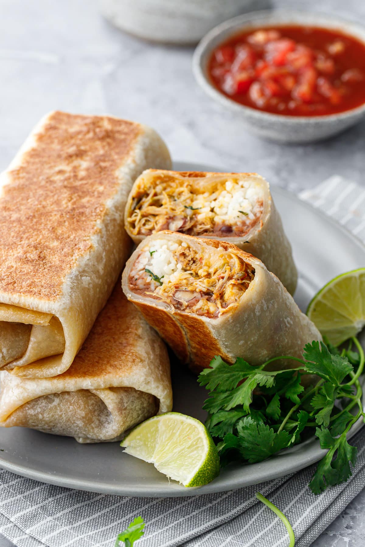 Crispy Chicken, Rice, & Bean Burritos on a gray plate, one burrito cut in half to show a cross section of the filling; fresh limes, cilantro, and a bowl of salsa on the side.