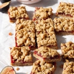 Squares of Fig, Apple & Vanilla Crumb Bars on a marble background, randomly arranged with a bowl of figs and a few cut figs scattered around.