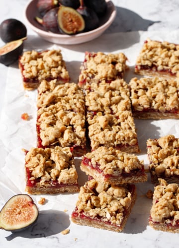 Squares of Fig, Apple & Vanilla Crumb Bars on a marble background, randomly arranged with a bowl of figs and a few cut figs scattered around.