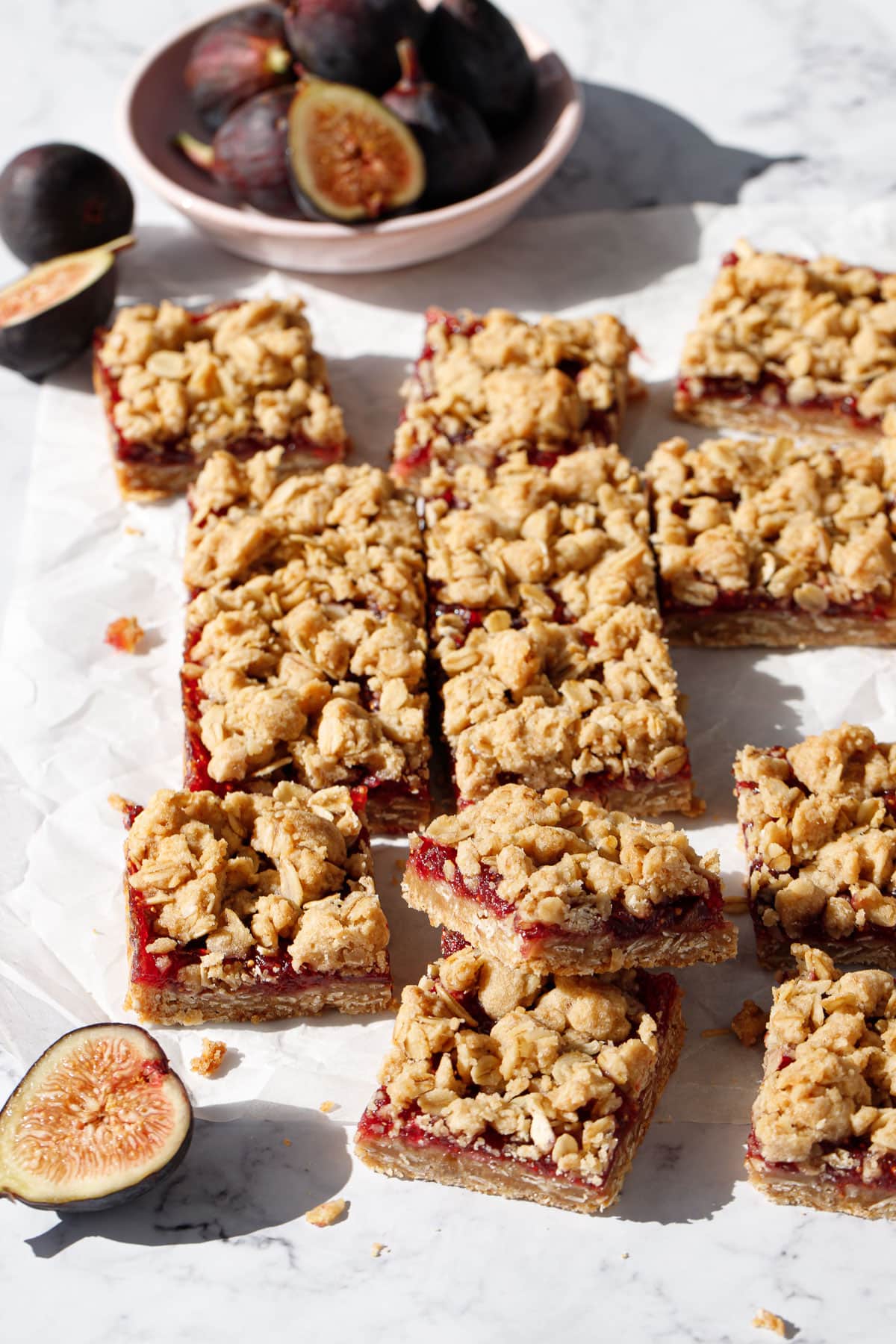 Squares of Fig, Apple & Vanilla Crumb Bars on a marble background, randomly arranged with a bowl of figs and a few cut figs scattered around.