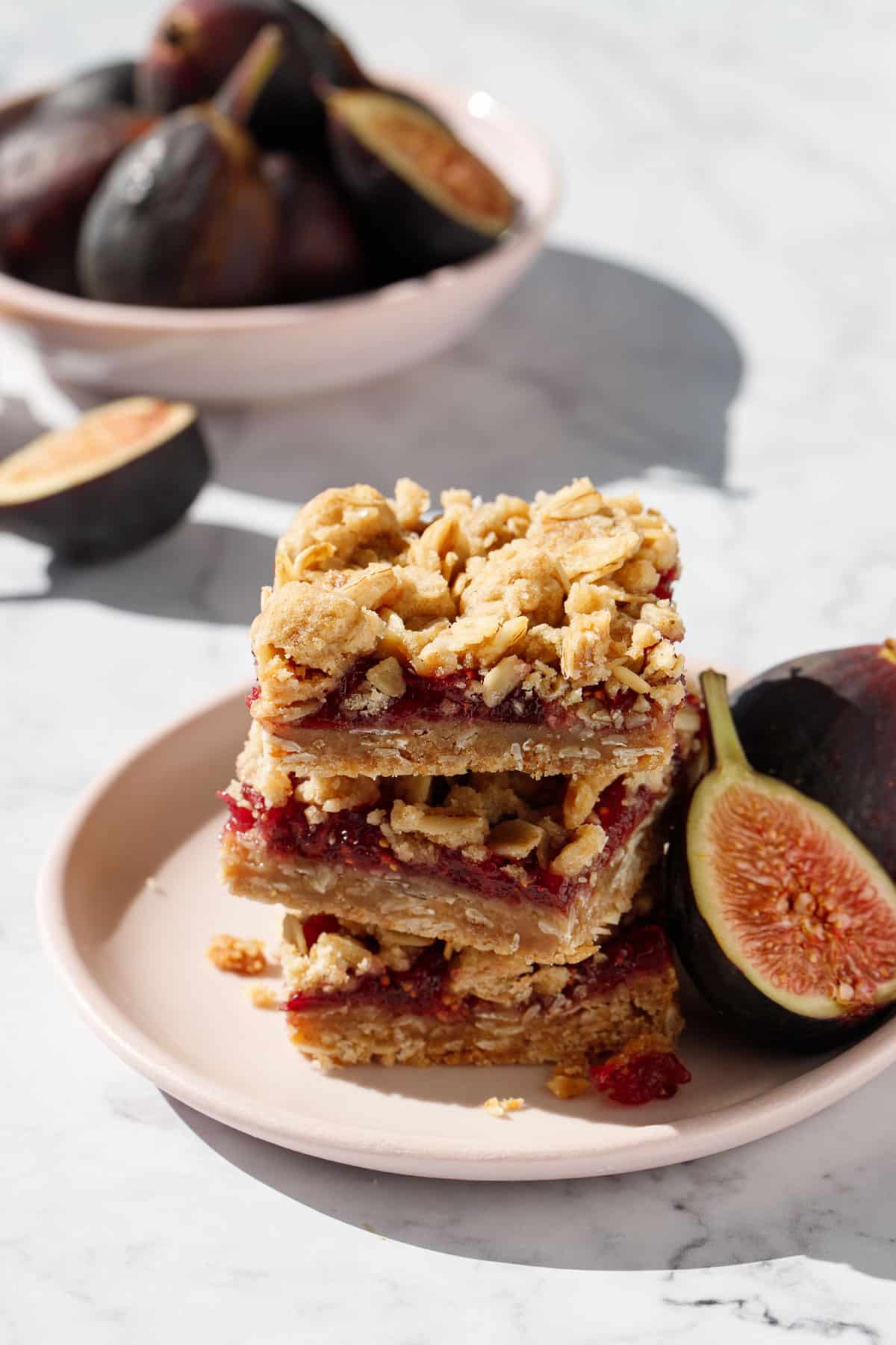Stack of three Fig, Apple & Vanilla Crumb Bars squares on a pink plate, with fresh figs and a bowl  of figs in the background.