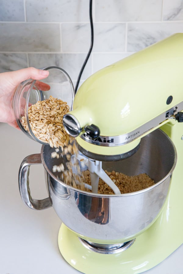 Pouring oats into the mixing bowl of a stand mixer fitted with the paddle attachment.