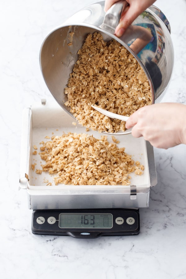 Pouring oat crumb mixture into a baking pan on a digital kitchen scale.