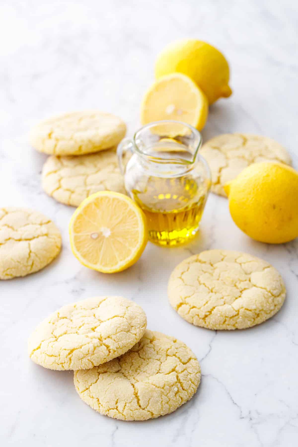 Lemon Olive Oil Sugar Cookies with crackly tops styled on a marble background with small glass pitcher of olive oil and whole and cut lemons.