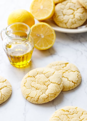 Crackly top Lemon Olive Oil Sugar Cookies on marble with a small glass pourer of olive oil, lemons, and a plate of more cookies in the background.