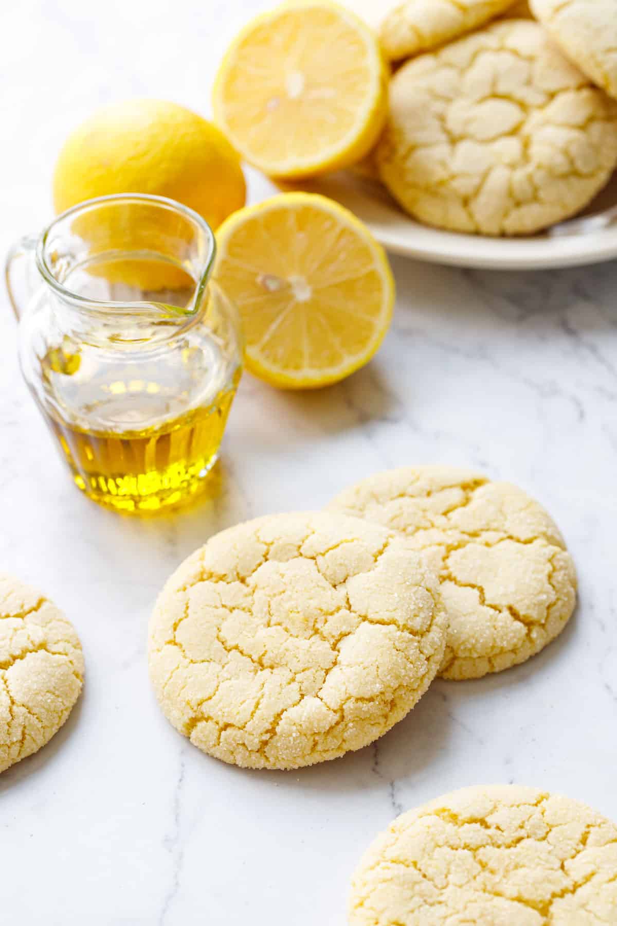 Crackly top Lemon Olive Oil Sugar Cookies on marble with a small glass pourer of olive oil, lemons, and a plate of more cookies in the background.