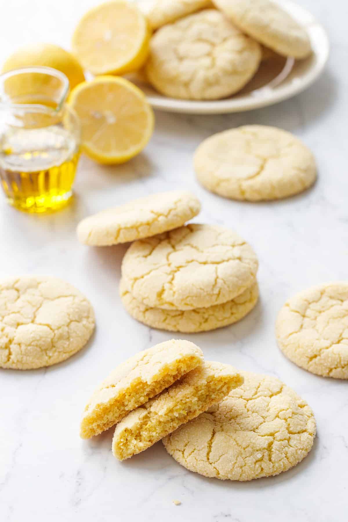 Scattered Lemon Olive Oil Sugar Cookies on a marble background with lemons, glass dish of olive oil and plate of cookies in the background.