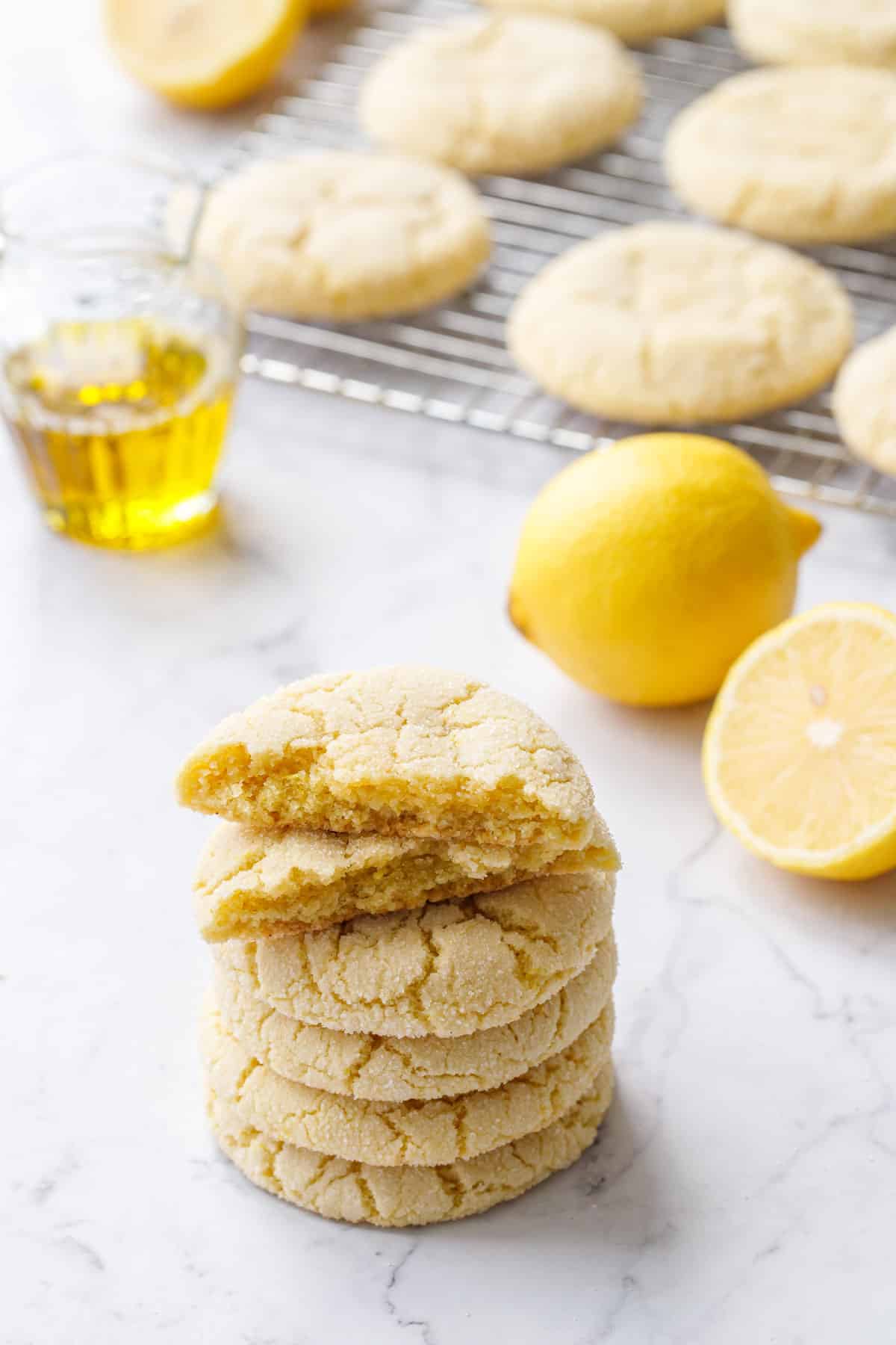 Stack of Lemon Olive Oil Sugar Cookies with the top cookie broken in half to show texture, rack of cookies plus lemons and a carafe of olive oil in the background.