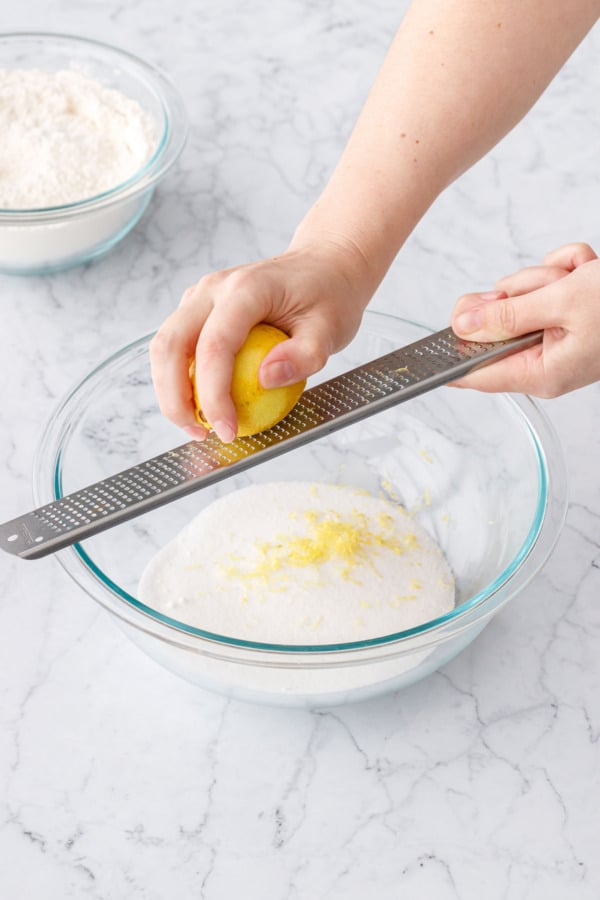 Hands zesting a lemon into a glass bowl filled with sugar.