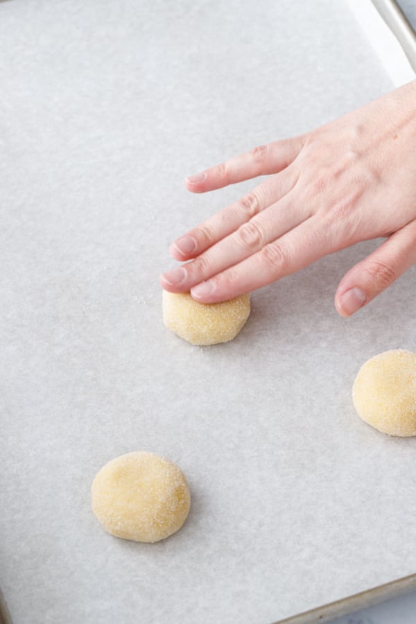 Hand pressing down on a ball of cookie dough on a parchment-lined cookie sheet to flatten it slightly.