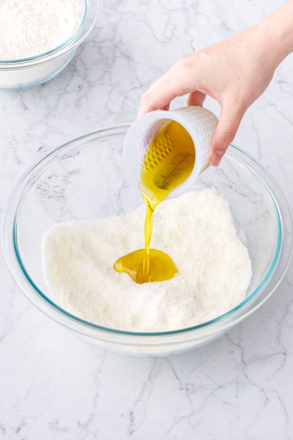 Pouring olive oil into a glass mixing bowl with lemon sugar.