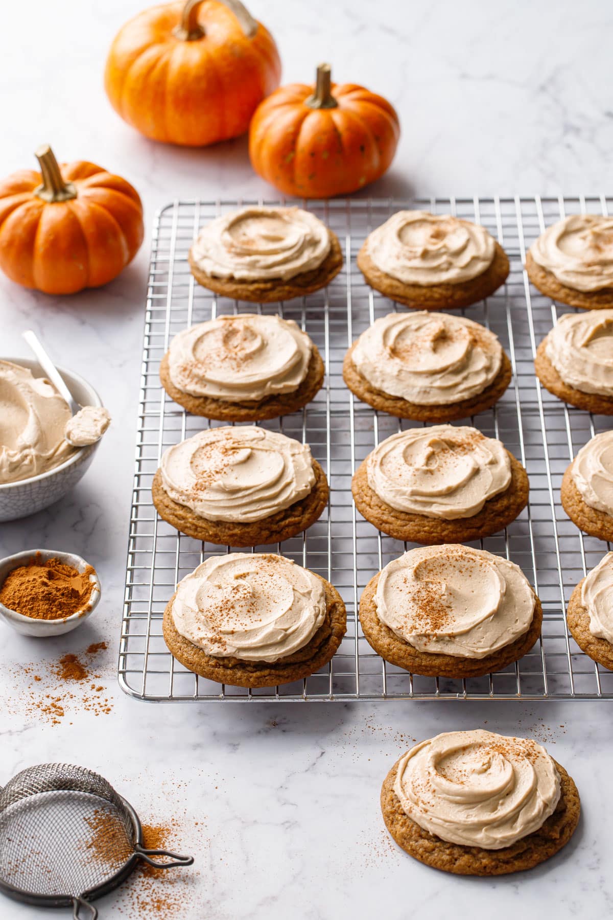 Rows of Caramel Pumpkin Cookies with Salted Caramel Frosting on a wire baking rack, with small pumpkins, bowl of frosting, and tiny bowl of cinnamon on the side.