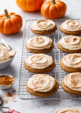 Wire baking rack with rows of frosted Caramel Pumpkin Cookies dusted with cinnamon, decorative orange pumpkins and bowl of frosting in the background.